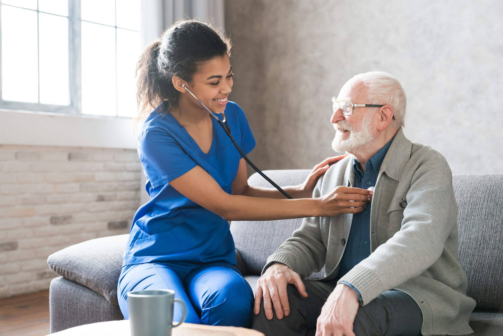 Female attending physician holding stethoscope listening old patient during homecare visit.